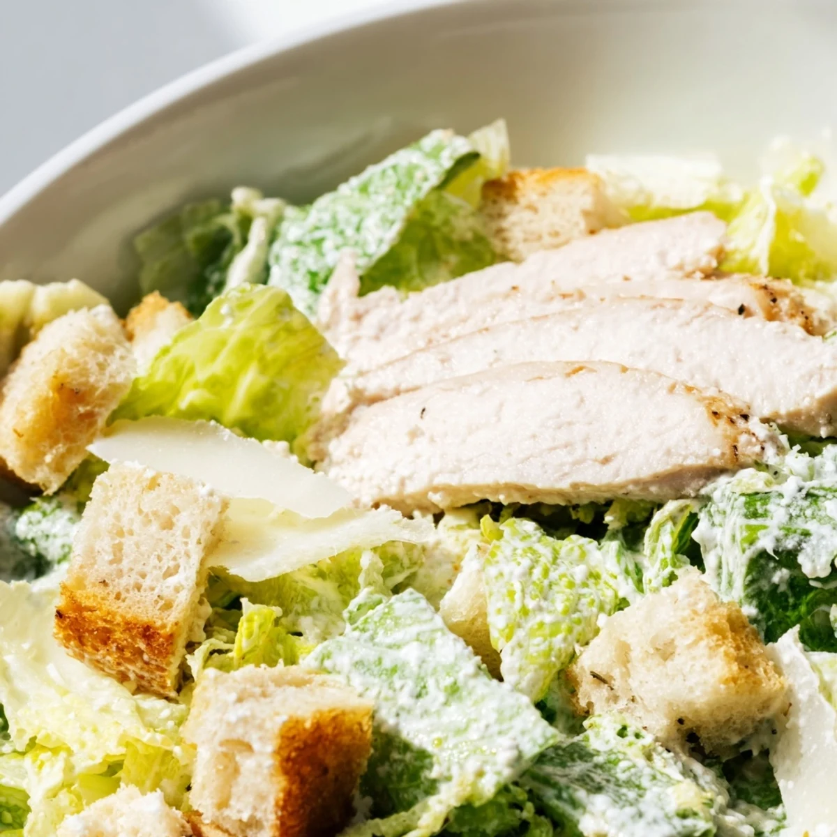 Sliced grilled chicken rests atop crisp romaine lettuce in this Chicken Caesar Salad, garnished with crunchy croutons and shaved Parmesan.