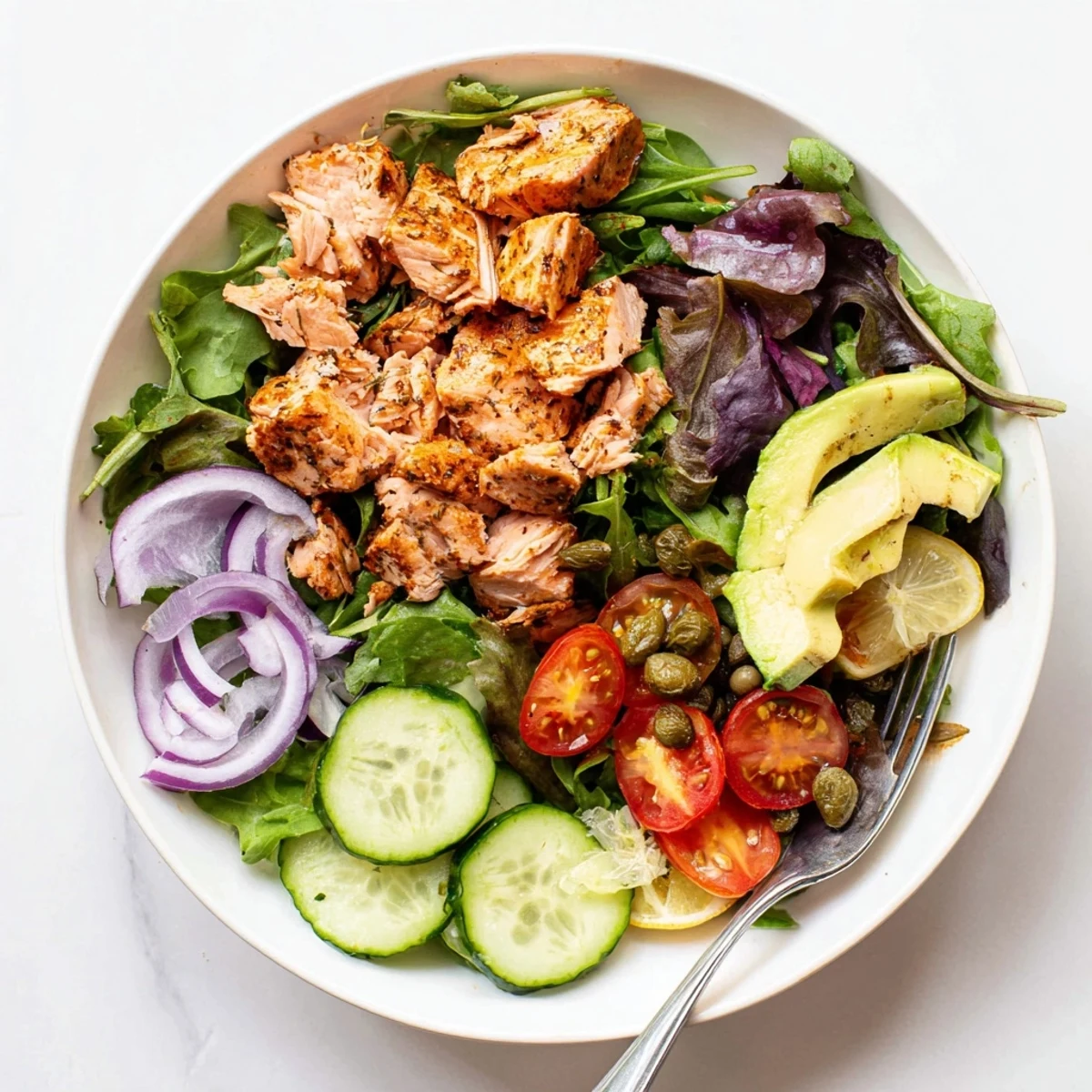 Overhead view of the Baked Salmon Lunch Salad in a white bowl, featuring crisp cucumbers, red onion, and capers beside tender baked salmon.