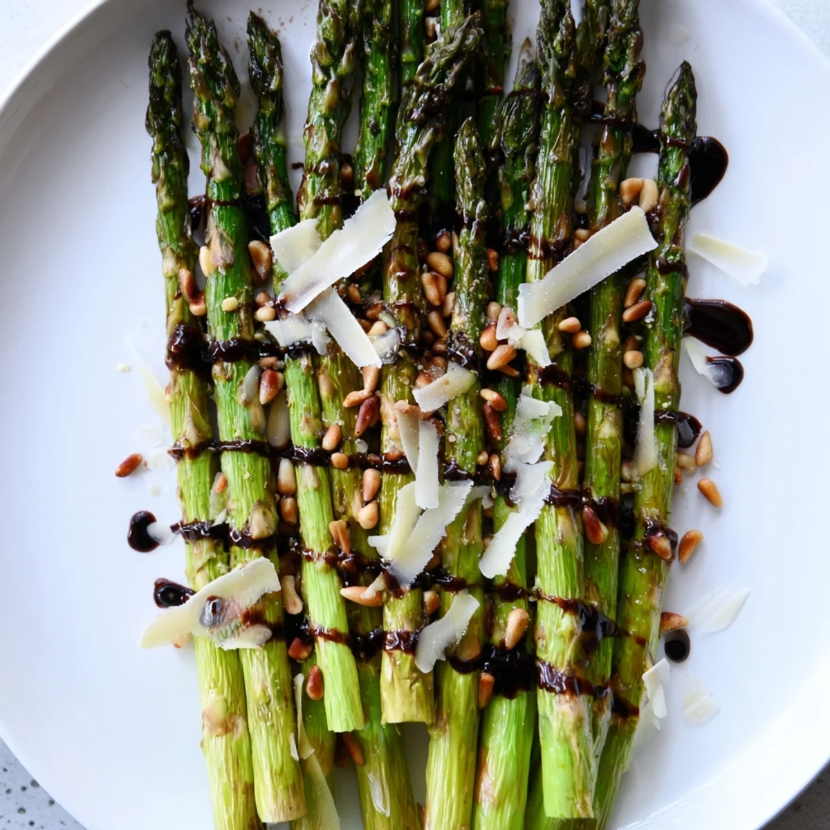 Fresh roasted asparagus with balsamic glaze on a white platter, garnished with pine nuts and Parmesan.
