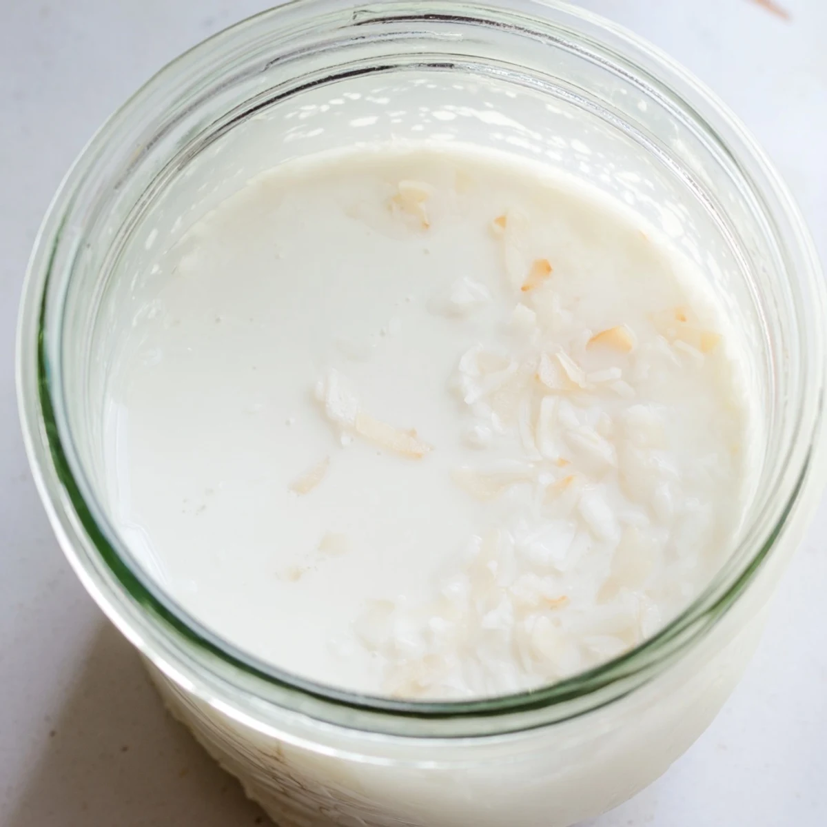Glass of creamy homemade coconut milk beside dried coconut flakes and a coconut on wooden surface